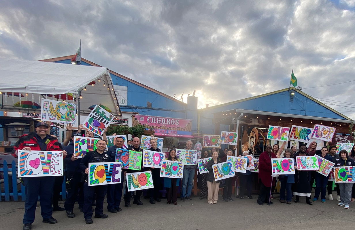 Group of people, including police, holding up colorful LOVE signs outside blue storefronts under a cloudy, bright sky.