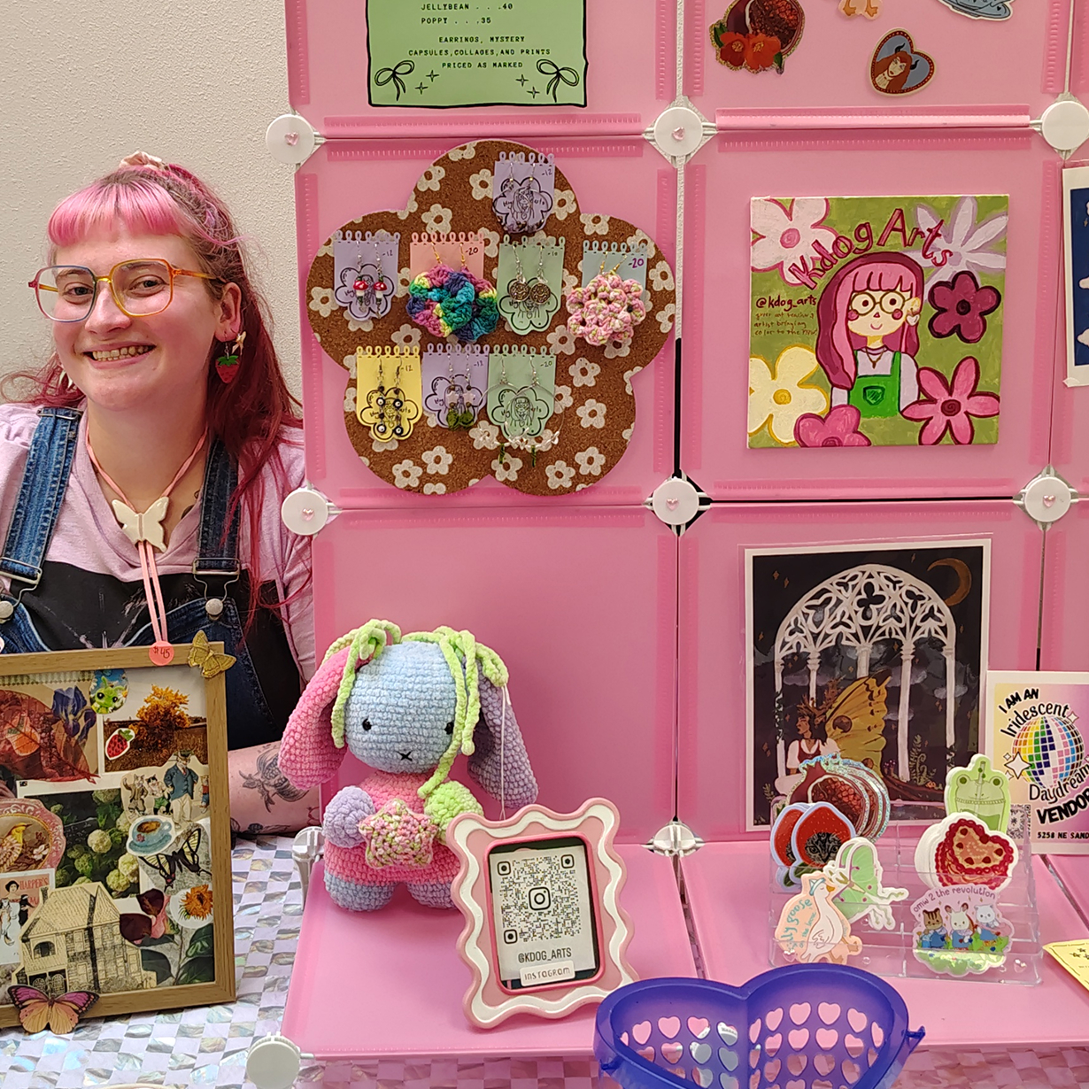 Smiling vendor with pink hair and glasses sits by a pink display featuring art, pins, a crocheted bunny, and stickers.