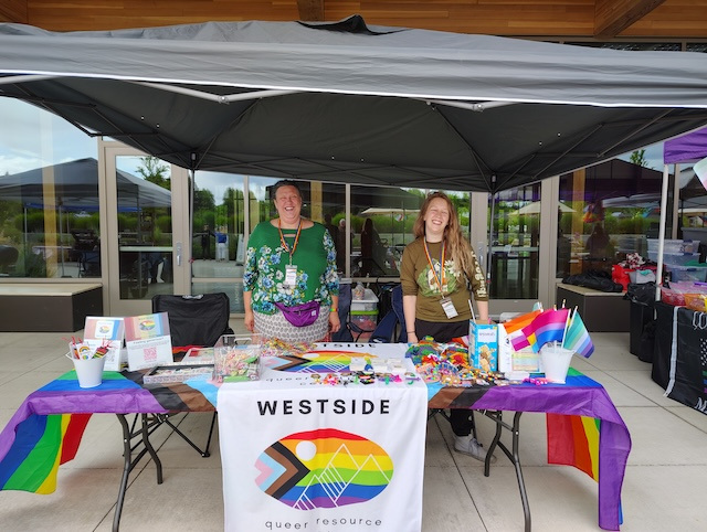 Two people smile behind a Pride-themed Westside Queer Resource booth with flags and colorful items at an outdoor event.